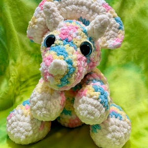Puede incluir: Un juguete de perro de ganchillo hecho a mano en colores pastel. El perro tiene grandes ojos azules, un hocico blanco y orejas blancas. El cuerpo es una mezcla de hilo rosa, amarillo, azul y blanco. El juguete está sobre un fondo verde.