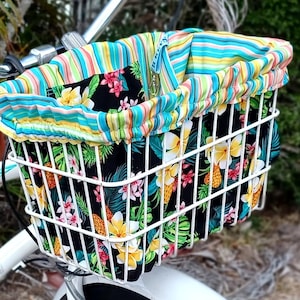 May include: A white wire bicycle basket featuring a fabric liner with a black background, yellow and white plumeria flowers, green palm leaves, and pineapples. The top edge has a ruffled trim in a multi-colored striped pattern of teal, yellow, and pink.