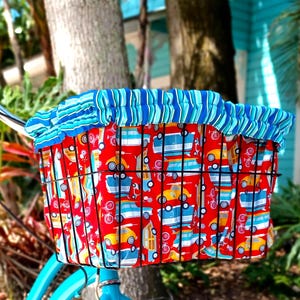 May include: A turquoise bicycle with a black wire basket and seat, both covered in a red fabric patterned with colorful cars. The basket has a blue and white striped fabric liner. The bike is parked outdoors.