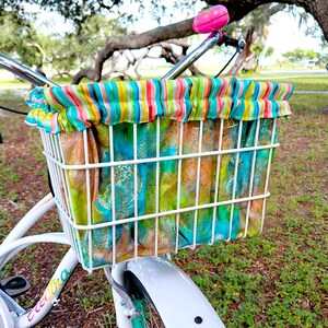 May include: A white wire bicycle basket featuring a removable fabric liner with a vibrant multicolored vertical stripe pattern at the top edge and a teal, green, and gold metallic floral print on the main body, mounted on white handlebars.