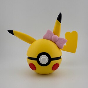May include: A yellow Pikachu-themed Pokeball with black-tipped ears and a yellow tail. The Pokeball has a black and white center, red circles, and a pink bow. The design is a playful take on the popular character.