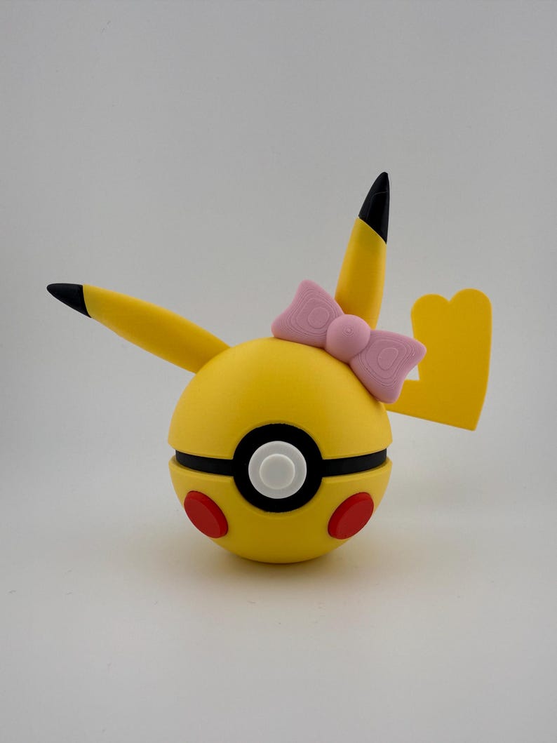 May include: A yellow Pikachu-themed Pokeball with black-tipped ears and a yellow tail. The Pokeball has a black and white center, red circles, and a pink bow. The design is a playful take on the popular character.