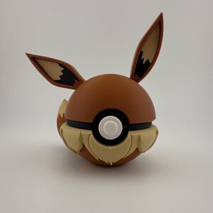 May include: A brown and tan Eevee-themed Pok&eacute; Ball. The ball features Eevee's ears, tail, and fur details. The centre has a white button surrounded by a black band. The ears are tan with brown tips.