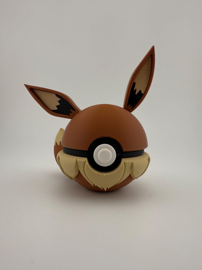 May include: A brown and tan Eevee-themed Pok&eacute; Ball. The ball features Eevee's ears, tail, and fur details. The centre has a white button surrounded by a black band. The ears are tan with brown tips.