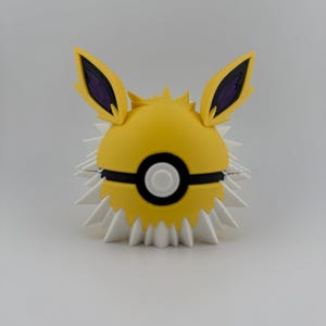 May include: A yellow and white Jolteon-themed Pok&eacute; Ball. The ball features a yellow sphere with black and white accents, including a black band and a white button. It has white spikes around the base and yellow ears with purple interiors.