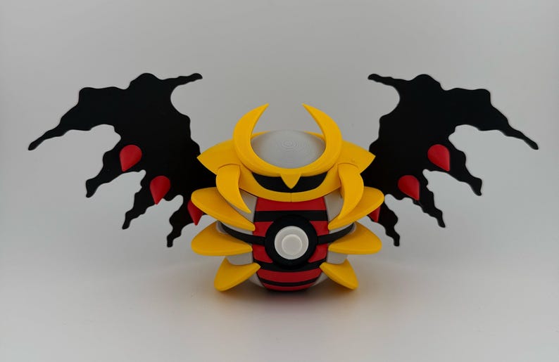 May include: A stylized, three-dimensional figure with a yellow helmet and body, black wings with red accents, and a red and black striped midsection. The figure has a white button-like detail in the center.