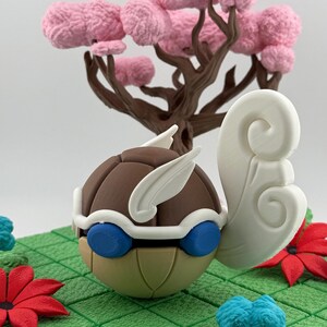 Pode incluir: Uma escultura artesanal e caprichosa com um personagem castanho, bege e azul com detalhes brancos e um grande elemento branco rodopiante. Uma &aacute;rvore castanha com flores cor-de-rosa ao fundo. A escultura assenta numa superf&iacute;cie verde texturizada com detalhes florais vermelhos e azuis.