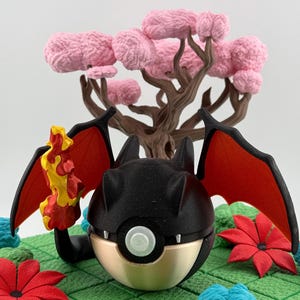 May include: A black and gold decorative figure with red wings and a flame. The figure is set on a green surface with red flowers and a pink tree in the background. The figure has a round shape with a white button.