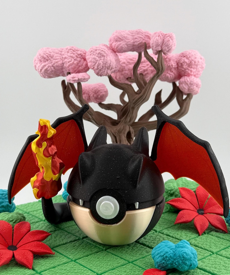 May include: A black and gold decorative figure with red wings and a flame. The figure is set on a green surface with red flowers and a pink tree in the background. The figure has a round shape with a white button.