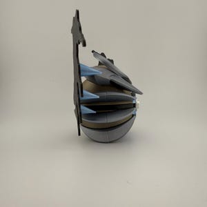 May include: A 3D-printed, futuristic-looking object with a spherical base and multiple angular, layered components. It features gray, tan, and blue accents, with a tall, black, decorative element extending upwards. The design suggests a mechanical or sci-fi theme.
