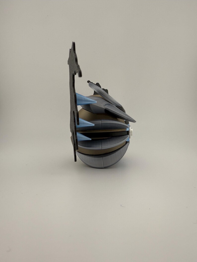 May include: A 3D-printed, futuristic-looking object with a spherical base and multiple angular, layered components. It features gray, tan, and blue accents, with a tall, black, decorative element extending upwards. The design suggests a mechanical or sci-fi theme.