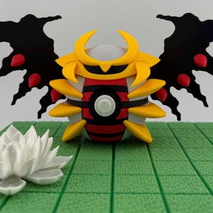 May include: A detailed, colorful figurine of a creature with yellow, red, black, and white accents. The figure has large black wings with red details and is positioned on a green, grid-patterned surface. A white lotus flower is in the foreground.