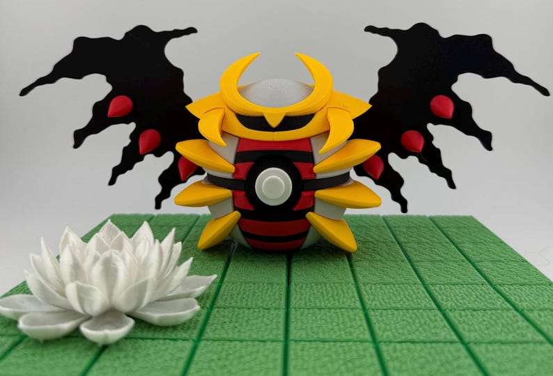 May include: A detailed, colorful figurine of a creature with yellow, red, black, and white accents. The figure has large black wings with red details and is positioned on a green, grid-patterned surface. A white lotus flower is in the foreground.