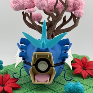 May include: A blue and turquoise 3D-printed figure of a serpentine creature with an open mouth, sharp teeth, and a fin. The figure is set against a backdrop of a pink tree and red flowers on a green surface.
