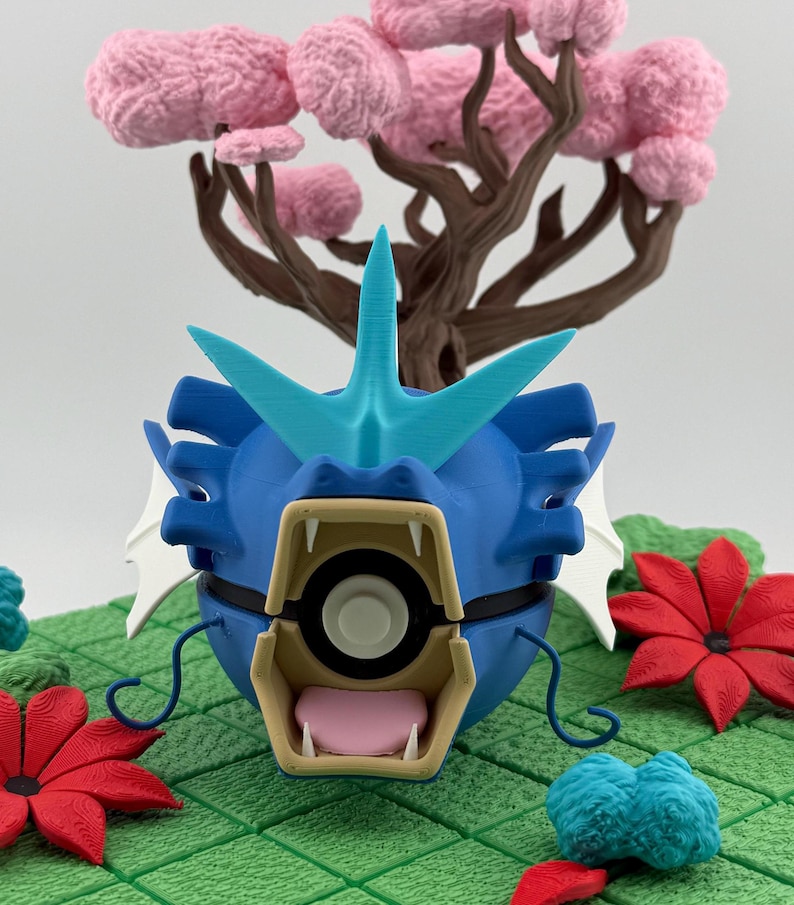May include: A blue and turquoise 3D-printed figure of a serpentine creature with an open mouth, sharp teeth, and a fin. The figure is set against a backdrop of a pink tree and red flowers on a green surface.