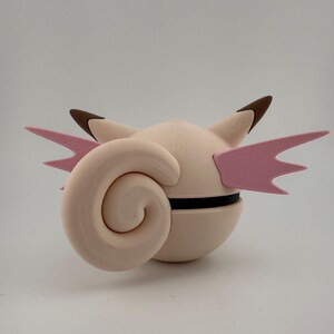 K&ouml;nnte beinhalten: Hellrosa, kugelf&ouml;rmige Figur mit einem gro&szlig;en, spiralf&ouml;rmigen Anhang und zwei rosa fl&uuml;gelartigen Strukturen. Die Figur hat braunspitzige Ohren und ein schwarzes Band um die Mitte. Das Design erinnert an eine Zeichentrickfigur.
