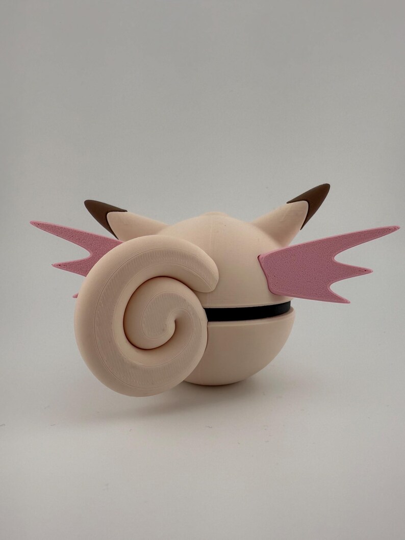 K&ouml;nnte beinhalten: Hellrosa, kugelf&ouml;rmige Figur mit einem gro&szlig;en, spiralf&ouml;rmigen Anhang und zwei rosa fl&uuml;gelartigen Strukturen. Die Figur hat braunspitzige Ohren und ein schwarzes Band um die Mitte. Das Design erinnert an eine Zeichentrickfigur.
