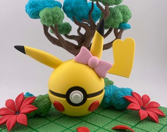 Op de afbeelding: Een gele Pikachu-sculptuur met een roze strik, rode wangen en zwartgepunte oren. De sculptuur staat tegen een achtergrond van een miniatuurboom met blauw en groen gebladerte, rode bloemen en een groene rasterbasis.