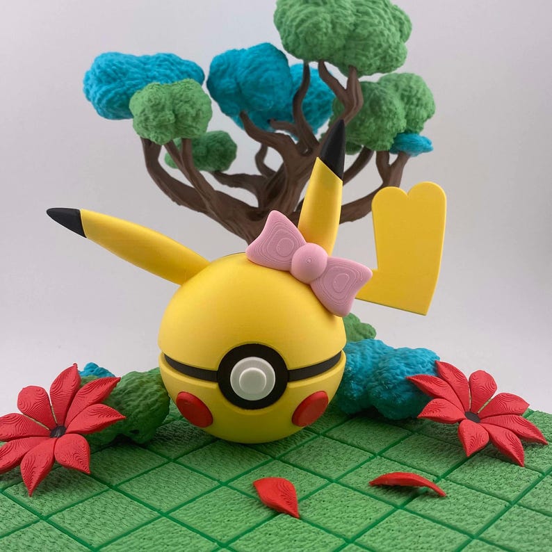 May include: A yellow Pikachu-themed sculpture with a pink bow, red cheeks, and black-tipped ears. The sculpture is set against a backdrop of a miniature tree with blue and green foliage, red flowers, and a green grid base.