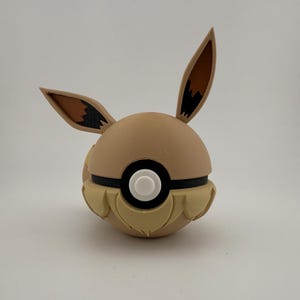 May include: A tan and brown Eevee-themed Pok&eacute; Ball. The ball features Eevee's ears, a black stripe, and a white button. The lower portion has a textured, fur-like design. A unique collectible for fans of the popular franchise.