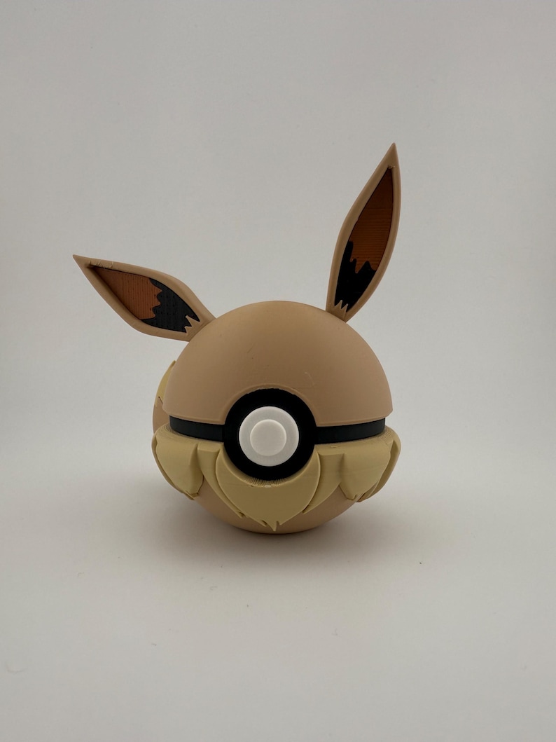 May include: A tan and brown Eevee-themed Pok&eacute; Ball. The ball features Eevee's ears, a black stripe, and a white button. The lower portion has a textured, fur-like design. A unique collectible for fans of the popular franchise.