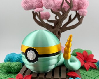 Op de afbeelding: Een 3D-geprint, bolvormig object in turquoise en goud, dat lijkt op een Poké Ball, staat op een miniatuur houten bankje. De scène bevat een bruine boom met roze bloesems, rode en blauwe bloemen en groen gebladerte.