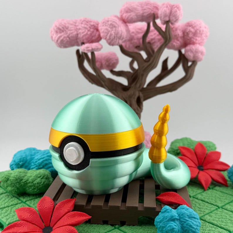 May include: A 3D-printed teal and gold ball-shaped object with a white button, resembling a Poke Ball, sits on a miniature wooden bench. The scene includes a brown tree with pink, fluffy blossoms, red and blue flowers, and green foliage.