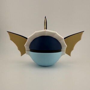 May include: A unique, stylized sculpture with a light blue, navy blue, and white color scheme. The sculpture has a rounded base, a segmented white top, and gold-colored wing-like elements on either side. A thin, black and gold element rises from the top.