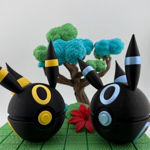 May include: Two black, spherical figures with yellow and blue accents, resembling cartoon characters, are displayed on a green, textured base. A miniature tree with blue and green foliage and a red flower are in the background.