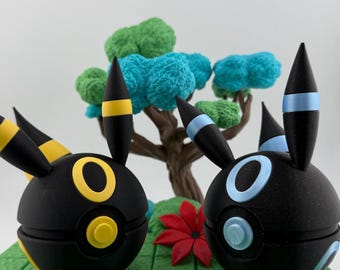 Op de afbeelding: Twee zwarte, bolvormige figuren met gele en blauwe accenten, die op stripfiguren lijken, worden weergegeven op een groene, getextureerde basis. Een miniatuurboom met blauw en groen gebladerte en een rode bloem staan op de achtergrond.