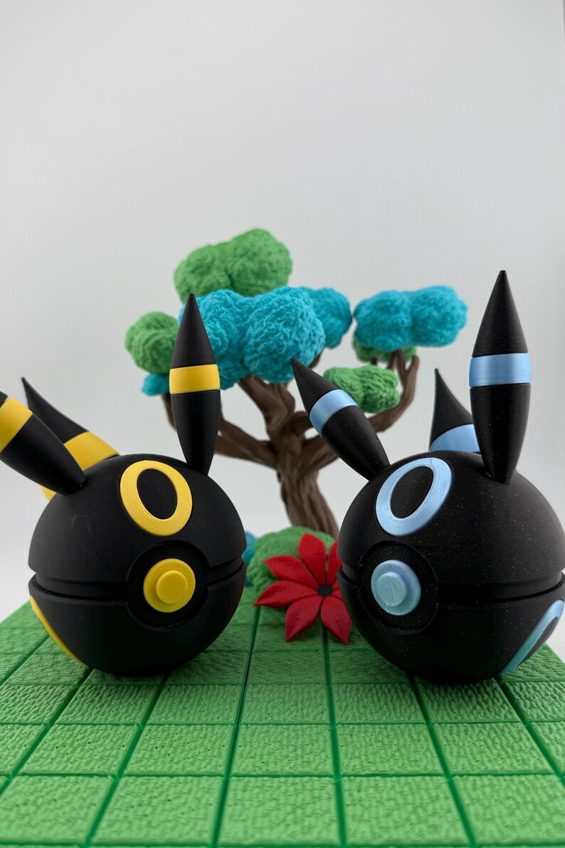 May include: Two black, spherical figures with yellow and blue accents, resembling cartoon characters, are displayed on a green, textured base. A miniature tree with blue and green foliage and a red flower are in the background.