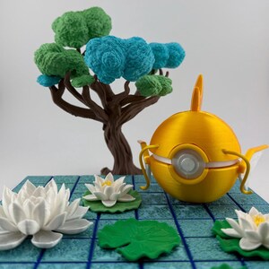 K&ouml;nnte beinhalten: Eine gelbe, kugelf&ouml;rmige Skulptur mit einer flossenartigen Struktur und einem zentralen, kreisf&ouml;rmigen Design. Sie steht auf einer blauen Fliesenbasis, zusammen mit wei&szlig;en Seerosen, gr&uuml;nen Seerosenbl&auml;ttern und einem Miniaturbaum mit gr&uuml;nem und blauem Laub.