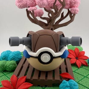 May include: A detailed, 3D-printed model of a Pokemon character, featuring a brown shell, gray cannons, and a cream-colored body. The model is set on a brown platform with red and blue floral accents, and a pink tree in the background.