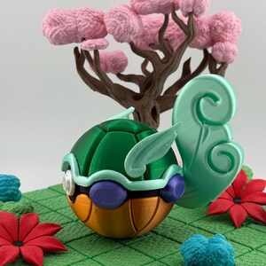 Pode incluir: Figura de tartaruga estilizada impressa em 3D, com carapa&ccedil;a verde, barriga laranja e detalhes roxos. Est&aacute; sobre uma superf&iacute;cie verde ladrilhada com detalhes florais vermelhos e azuis, e uma &aacute;rvore castanha com flores cor-de-rosa ao fundo.