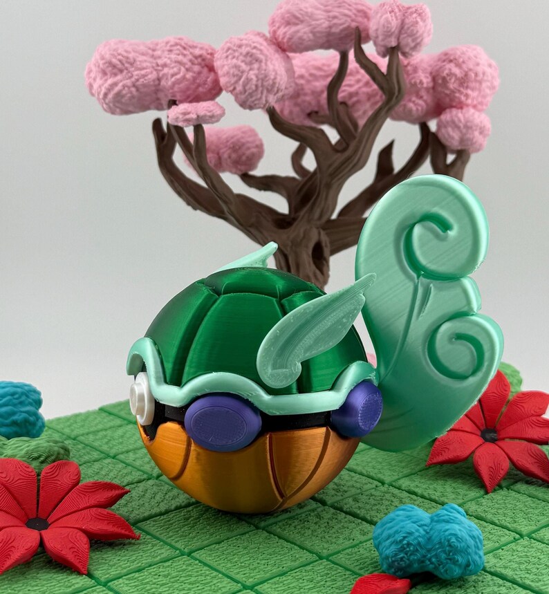 Pode incluir: Figura de tartaruga estilizada impressa em 3D, com carapa&ccedil;a verde, barriga laranja e detalhes roxos. Est&aacute; sobre uma superf&iacute;cie verde ladrilhada com detalhes florais vermelhos e azuis, e uma &aacute;rvore castanha com flores cor-de-rosa ao fundo.