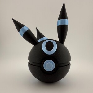 May include: A black spherical figurine with blue accents, featuring three pointed projections and circular details. The design includes a light blue band around the projections and light blue circles on the sphere's surface. The figurine is set against a plain background.