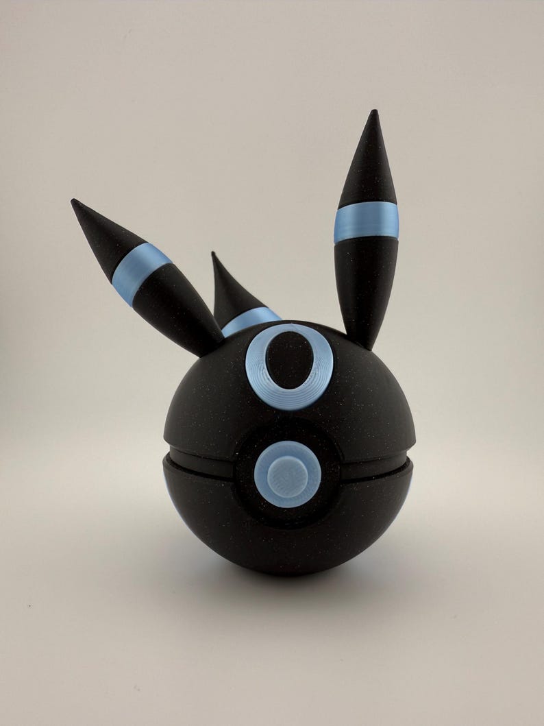 May include: A black spherical figurine with blue accents, featuring three pointed projections and circular details. The design includes a light blue band around the projections and light blue circles on the sphere's surface. The figurine is set against a plain background.