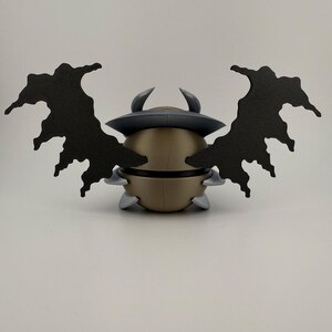 May include: A gold-colored, spherical figurine with a dark gray hat and two curved horns. It has two large, black, wing-like structures extending outwards. The figurine also has small, gray, triangular protrusions around its base.