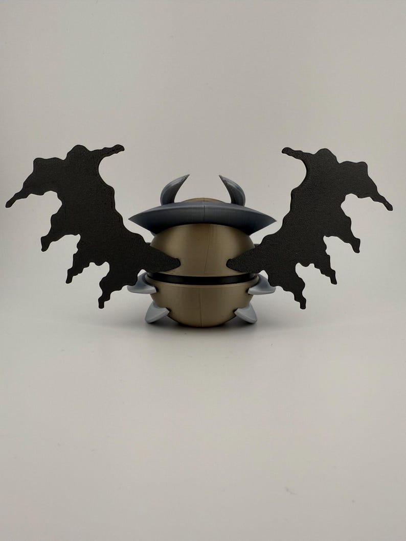 May include: A gold-colored, spherical figurine with a dark gray hat and two curved horns. It has two large, black, wing-like structures extending outwards. The figurine also has small, gray, triangular protrusions around its base.