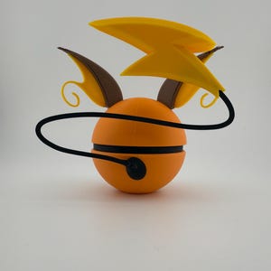 May include: An orange, spherical figurine with black stripes and a black cord. It features yellow lightning bolt-shaped accents and brown ear-like protrusions. The design is reminiscent of a popular animated character.