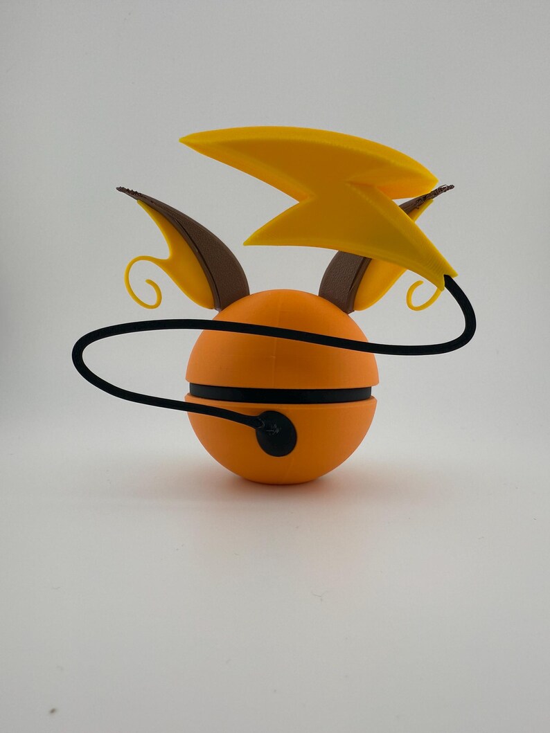 May include: An orange, spherical figurine with black stripes and a black cord. It features yellow lightning bolt-shaped accents and brown ear-like protrusions. The design is reminiscent of a popular animated character.