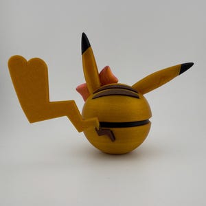 May include: A yellow, spherical figurine with black stripes and a lightning bolt tail. The figurine has two pointed ears with black tips and a small pink bow. The overall design is reminiscent of a popular cartoon character.
