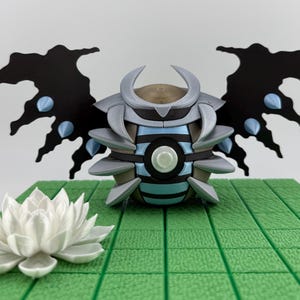 May include: A detailed 3D-printed sculpture of a stylized insect-like creature with black wings and blue accents. The sculpture is set on a green tiled surface, with a white lotus flower in the foreground.