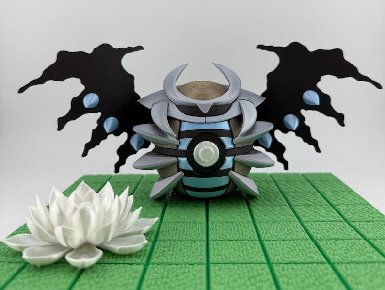 May include: A detailed 3D-printed sculpture of a stylized insect-like creature with black wings and blue accents. The sculpture is set on a green tiled surface, with a white lotus flower in the foreground.
