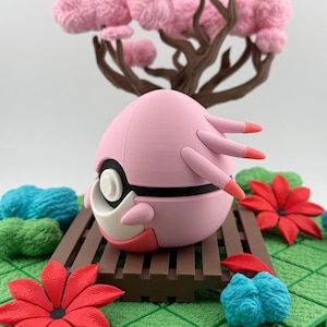 May include: A pink, spherical figurine with black and white accents, and orange-tipped appendages. It sits on a brown platform with a miniature tree with pink, fluffy foliage in the background. Red and blue floral accents are also present.