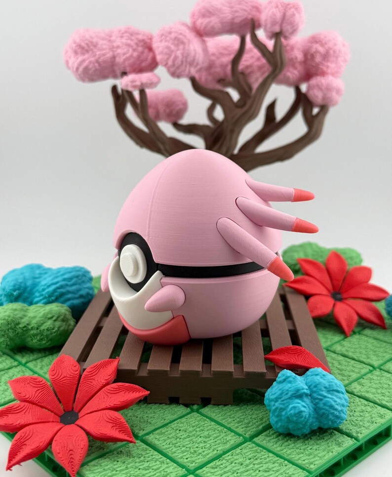 May include: A pink, spherical figurine with black and white accents, and orange-tipped appendages. It sits on a brown platform with a miniature tree with pink, fluffy foliage in the background. Red and blue floral accents are also present.