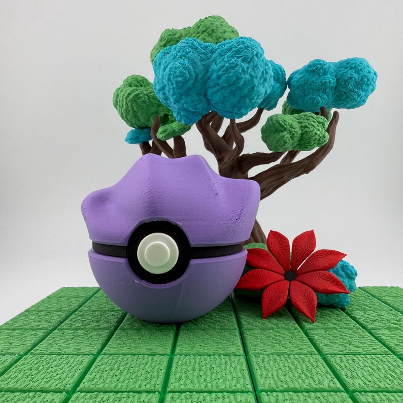 May include: A purple and black decorative ball with a white button, resembling a Poke Ball, sits in front of a miniature tree with blue and green foliage. A red flower is to the right. The scene is set on a green, textured surface.