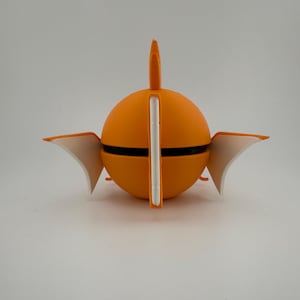 K&ouml;nnte beinhalten: Ein leuchtend orangefarbenes, kugelf&ouml;rmiges Objekt mit einem schwarzen Band, wei&szlig;en Akzenten und fl&uuml;gelf&ouml;rmigen Elementen. Ein vertikaler wei&szlig;er Balken teilt die Kugel, mit einer flossenartigen Struktur oben. Das Objekt steht vor einem schlichten wei&szlig;en Hintergrund.