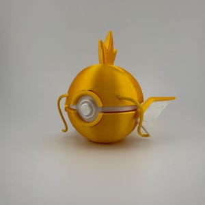 K&ouml;nnte beinhalten: Ein gelbes, kugelf&ouml;rmiges Objekt, 3D-gedruckt, das einem Fisch &auml;hnelt. Es hat eine Flosse, ein kronenartiges Element und ein silberfarbenes kreisf&ouml;rmiges Detail. Das Objekt befindet sich vor einem schlichten, hellen Hintergrund.
