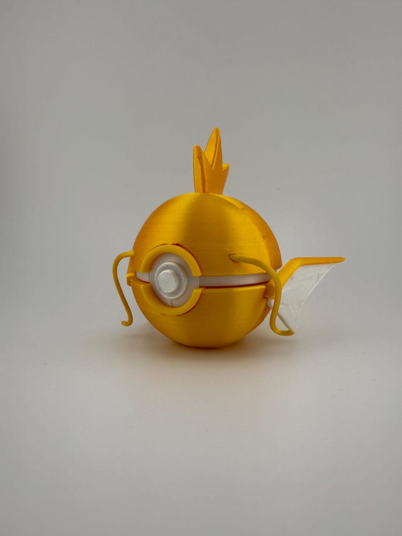 K&ouml;nnte beinhalten: Ein gelbes, kugelf&ouml;rmiges Objekt, 3D-gedruckt, das einem Fisch &auml;hnelt. Es hat eine Flosse, ein kronenartiges Element und ein silberfarbenes kreisf&ouml;rmiges Detail. Das Objekt befindet sich vor einem schlichten, hellen Hintergrund.